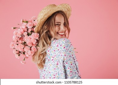 Close up portrait of a lovely young woman in summer dress and straw hat holding carnations bouquet and looking over her shoulder isolated over pink background