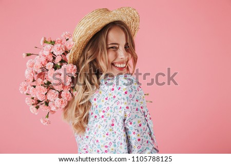 Close up portrait of a lovely young woman in summer dress and straw hat holding carnations bouquet and looking over her shoulder isolated over pink background