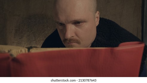 Close Up Of A Portrait Of Man Reading A Paper Book. He Is Sitting On Porch Of A Village House. The Concept Of Reading Paper Books, Studying And Hobbies. Dark Night. Old, Retro Decor Burlap Background