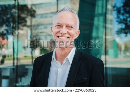 Close up portrait of mature adult business man with gray hair and suit smiling and looking at camera with succesful attitude. Happy corporate lawyer with white perfect teeth standing at workspace