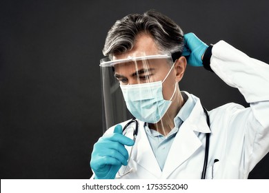 Close Up Portrait Of Mature Male Doctor In Medical Mask And Blue Gloves Wearing Face Shield Or Protective Gear While Standing Against Dark Background. Healthcare. Coronavirus, Covid-19. Lockdown