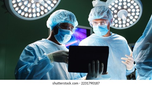 Close up portrait of mixed-races doctors male surgeons speaking and typing on tablet while standing in operating room performing operation on patient under the bright lamps in hospital during surgery - Powered by Shutterstock