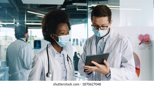 Close up portrait of mixed-races healthcare workers doctors in medical masks standing in hospital cabinet discussing health problem and tapping on tablet device. Professional physicians in clinic - Shutterstock ID 1870627048