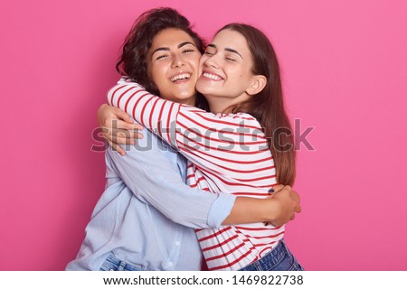 Close up portrait off positive ladies, frienads or sisters standing next to each other, having warm hug, posing with pleasant smiles, isolated over pink background. Friendship and happyness concept.