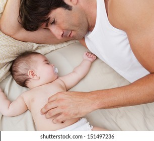 Close Up Portrait Over Head View Of A Father And His Baby Daughter Relaxing On A Bed At Home, With The Infant Sleeping And Dad Caring For Her.