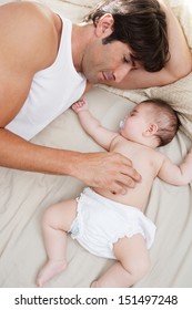 Close Up Portrait Over Head View Of A Father And His Baby Daughter Relaxing On A Bed At Home, With The Infant Sleeping And Dad Caring For Her.