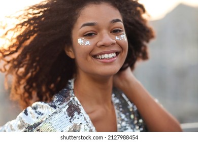 Close up portrait of overjoyed charming african american woman with curly hair and glitter on cheekbones smiling happily at camera while moving dancing outdoors, festive atmosphere in the air - Shutterstock ID 2127988454