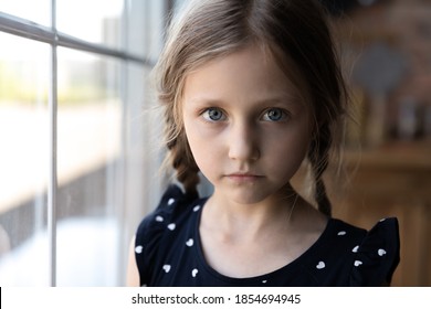 Close Up Portrait Of Pensive Serious Little School Preschool Age Girl Standing By Window At Home Looking At Camera With Sad Unhappy Face Having Problem Trouble Thinking About Bad Relations In Family.