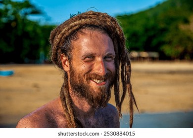 Close up portrait photography of a male model with stylish beard and dreadlocks hairstyle with happy smiling face expression standing at the tropical sandy beach in beautiful natural environment.  - Powered by Shutterstock