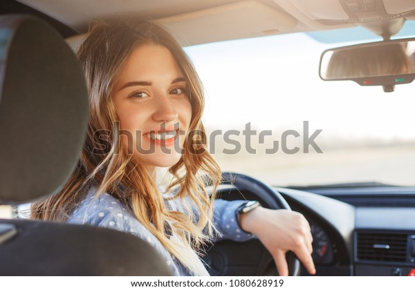 Close up portrait of pleasant looking female with\
glad positive expression, being satisfied with unforgettable\
journey by car, sits on driver`s seat, enjoys music. People,\
driving, transport\
concept