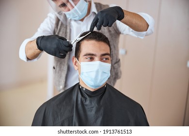 Close Up Portrait Of Pleasure Client Wearing Black Peignoir Sitting In Medicine Mask And Looking Away While Barber Cutting His Hair In Beauty Salon