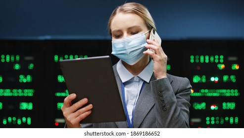 Close up portrait of pretty female system administrator in mask standing in data storage room working on digital security of processors and typing on tablet while talking on cellphone. Big data