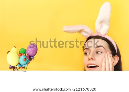 Close up portrait of pretty impressed young caucasian woman wear bunny fluffy ears, shocked fun looking at easter colorful eggs, posing isolated over yellow color background in studio with copy space