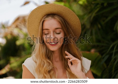  Close up portrait of Pretty traveling   blond woman in straw hat posing  in trpical stylish hotel. White summer clothes. Perfect wavy hairs. 
