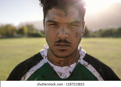 Close Up Portrait Of Rugby Player With Muddy Face And Uniform On Field. Young Sportsman Staring At Camera On Ground.