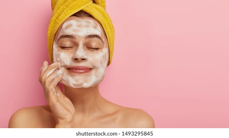 Close up portrait of satisfied beautiful woman massages face, washes with soap, poses bare shouldes, has healthy smooth skin, keeps eyes closed, isolated over pink background free space for your promo