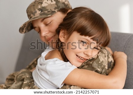 Close up portrait of satisfied little girl with dark hair and braids with mother, soldier woman wearing camouflage uniform and cap posing with her daughter after returning home from war.