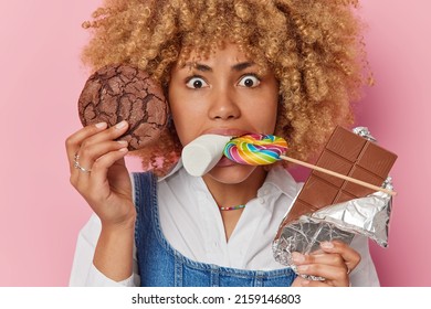 Close up portrait of serious curly haired young woman eats various sweets has mouth full of harmful food focused into camera wears white shirt isolated over pink background. Sweet life concept