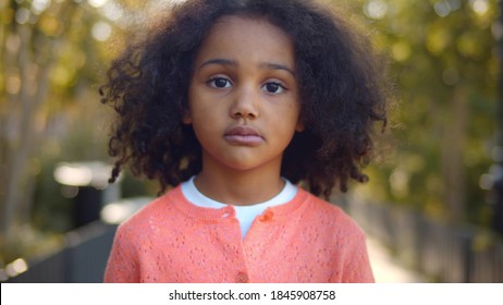 Close Up Portrait Of Serious Little African Girl Looking At Camera. Portrait Of Adorable Preschool Afro-american Kid Standing In Ark Over Blurred Background