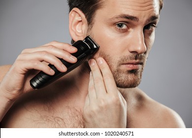 Close Up Portrait Of A Serious Shirtless Man Shaving Beard With An Electric Razor Isolated Over Gray Background