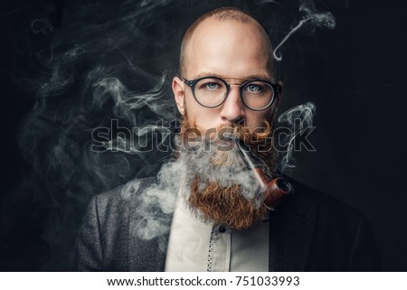 Close up portrait of shaved head aristocratic male in eyeglasses smoking pipe over grey background.