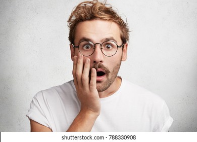 Close up portrait of shocked frightened bearded young male stares at camera through round spectacles, keeps mouth widely opened, covers mourh with hand, isolated over white concrete background
