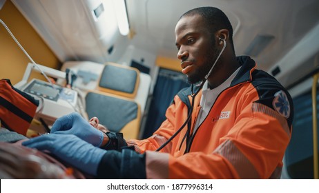 Close Up Portrait Shot Of An African American Professional Paramedic Providing Medical Help To An Injured Patient On The Way To Hospital. Emergency Care Assistant Using Stethoscope In An Ambulance.