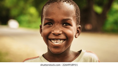 Close Up Portrait of a Shy Authentic African Boy Smiling at the Camera with a Blurred Rural Village in the Background. Black Male Kid Representing Future, Hope, and Acceptance. Documentary Footage - Shutterstock ID 2264918911