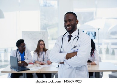 Close Up Portrait Of Smiling Afro American Male Doctor. Medical Assistant Or Student In White Uniform With Stethoscope. Handsome Male Doctor With A Medical Group At Meeting Room.