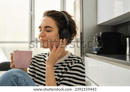 Close up portrait of smiling brunette woman, student in headphones, listens music, drinks cup of tea in morning, sits in kitchen and enjoys her daily routine.