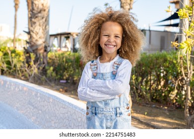 Close Up Portrait Of A Smiling Child Girl In Spring Park On Sunny Warm Day With Arms Folded.