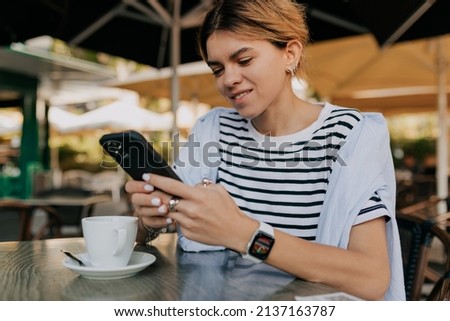 Close up portrait of smiling European woman in striped t-shirt in smart watch is using smartphone and drinking morning coffee in outdoor cafe. High quality photo
