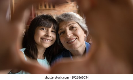 Close Up Portrait Of Smiling Mature Hispanic Grandmother And Little 7s Granddaughter Have Fun Make Selfie Together. Happy Senior Latino Grandparent And Small Girl Child Make Heart Love Hand Gesture.