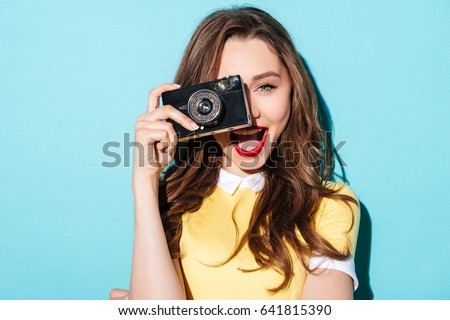 Close up portrait of a smiling pretty girl in dress taking photo on a retro camera isolated over blue background