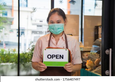 Close Up Portrait Smiling Young Asian Woman In A Beige Shirt With A Apron Wearing A Face Mask Holding An Open Sign Board In Front Of Coffee Shop Door, New Normal Concept.