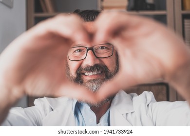 Close Up Portrait Of Smiling Young Caucasian Male Nurse Or GP In White Medical Uniform Show Heart Love Hand Gesture. Happy Man Doctor Show Support And Care To Patients Or Client In Hospital.
