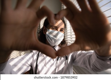 Close Up Portrait Of Smiling Young Female Doctor In White Medical Uniform And Facemask Show Heart Love Hand Gesture. Happy Woman GP In Facemask Show Support, Empathy And Care. Corona Concept.