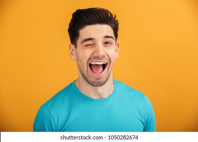 Close up portrait of a smiling young man in t-shirt looking at camera and winking isolated over yellow background