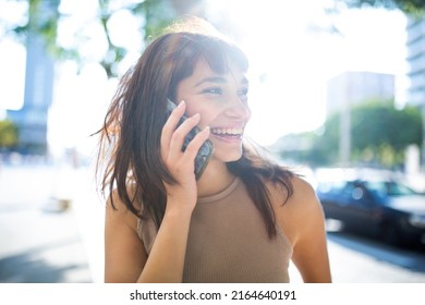 Close Up Portrait Of Smiling Young Woman Talking On Phone Outside In City With Sunshine From Behind
