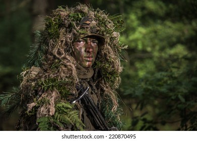 Close Portrait Of The Soldier Wearing Ghillie Suit, Face Painted With Camouflage Paints