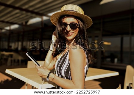 Close up portrait of spectacular woman with dark dark, wearing hat, sunglasses and summer t-shirt posing at camera with smartphone 