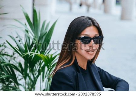 Close up portrait of stylish luxury dressed female model in sunglasses sitting outside against green plants. Tanned successful woman relaxing at outside cafe. Wealthy lifestyle. 