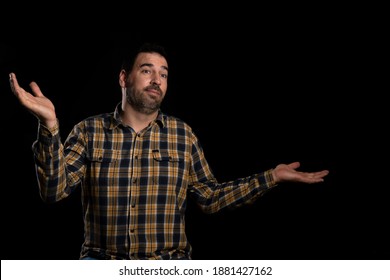 Close Up Portrait Of A Surprised Young Man In Yellow Plaid Shirt Posing Isolated On Black Studio Background, Raising Palms Of Hands And Squeezing Shoulders, Handsome Unshaven Man Not Looking At Camera