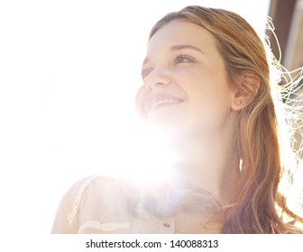 Close up portrait of a teenage girl in a city street during a sunny day, looking away from the camera and smiling against the sky with sun rays filtering through her neck.