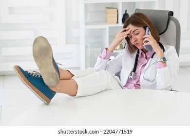 Close Up Portrait Of Tired Female Doctor Sitting At The Desktop And Talking To Phone In The Office Of Modern Clinic