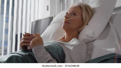 Close Up Portrait Of Tired Sick Woman Lying In Hospital Bed And Drinking Water. Aged Female Patient With Breathing Tube Holding Cup With Beverage Resting In Ward