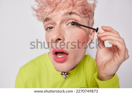 Close up portrait of transgender guy puts on makeup. Young gay man applies mascara on eyelashes keeps mouth opened cares about his appearance wears green jumper isolated over white background Foto stock © 