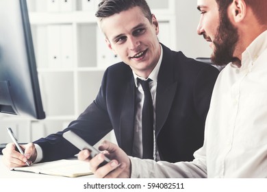 Close Up Portrait Of Two Business Partrners At Their Workplace. One Is Holding His Cell Phone, The Second Is Writing And Looking At Him. Toned Image