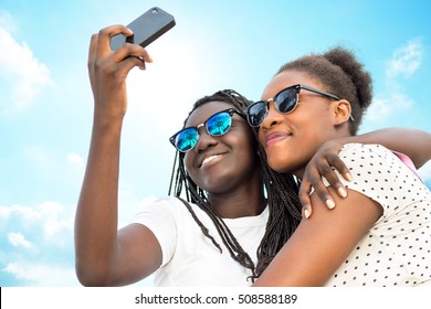 Close up portrait of two diverse african teen girls wearing sun glasses taking self portrait with phone against blue sky. - Powered by Shutterstock