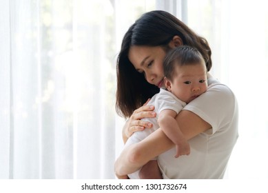 Close Up Portrait View Of Beautiful Asian Mother Holding Her Newborn Baby In Her Arms And Support Baby's Back By Her Hand With White Curtain In Background. She Is Feeling Happy To Stay With Her Child.